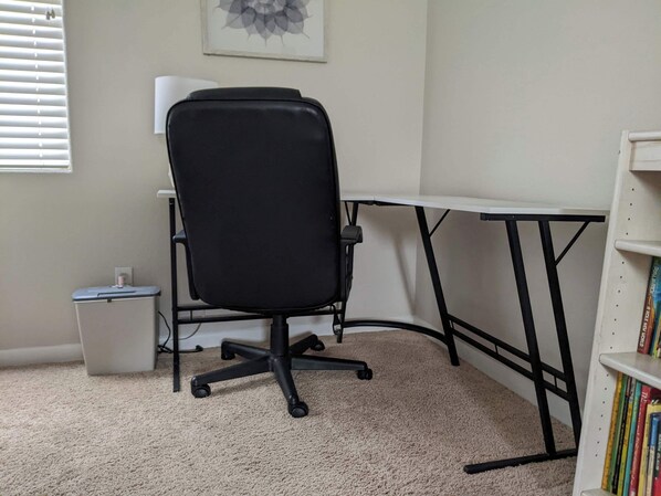 Una habitación con una silla de oficina negra, un escritorio blanco, una estantería y suelo alfombrado.