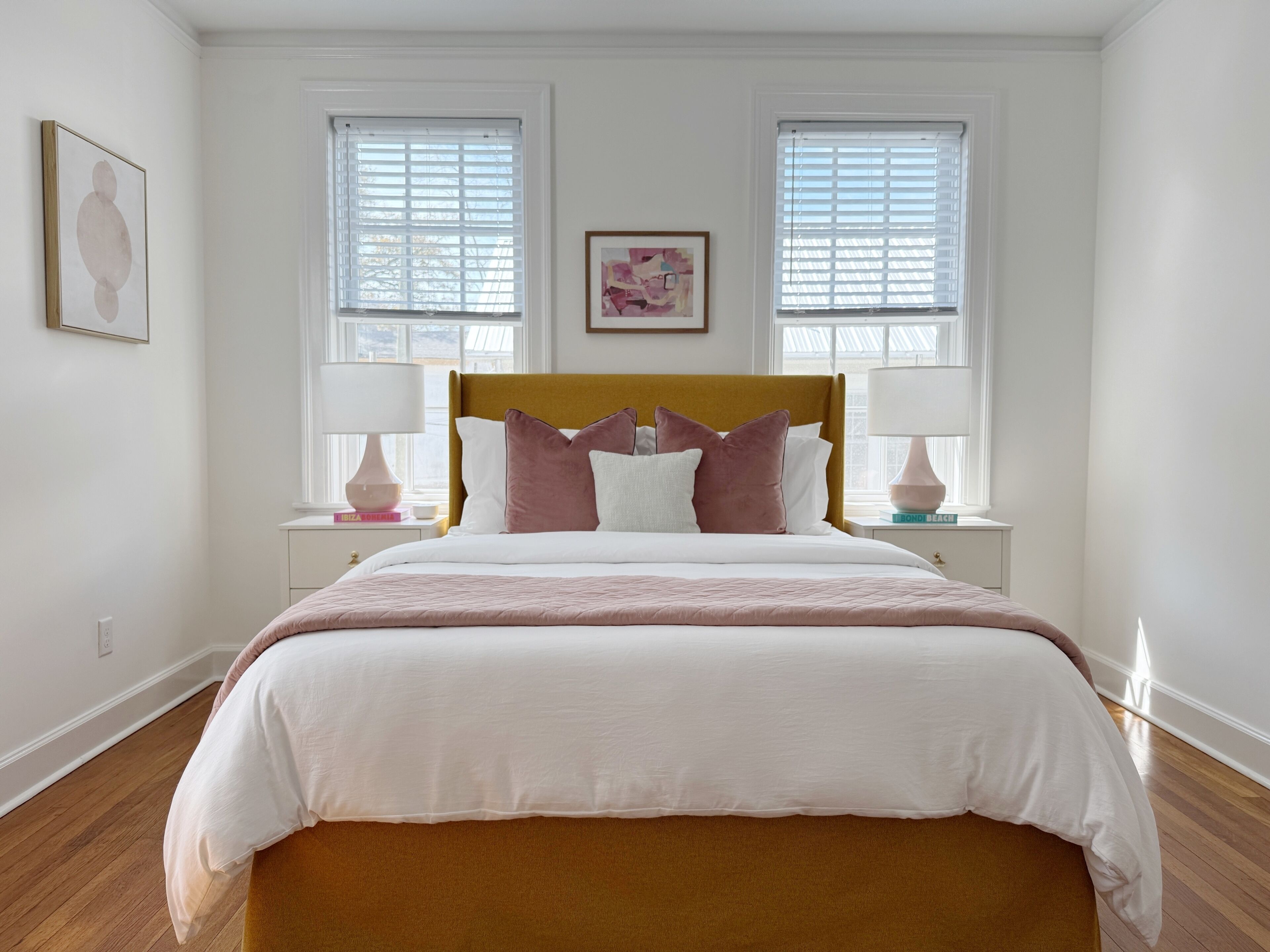 A bedroom with a large bed, two bedside tables with lamps, and two windows with blinds.