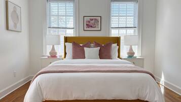 A bedroom with a large bed, two bedside tables with lamps, and two windows with blinds.