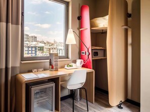 Une chambre d’hôtel compacte comprenant un bureau, une chaise, un mini-réfrigérateur, un meuble de rangement rouge et une vue sur le paysage urbain.