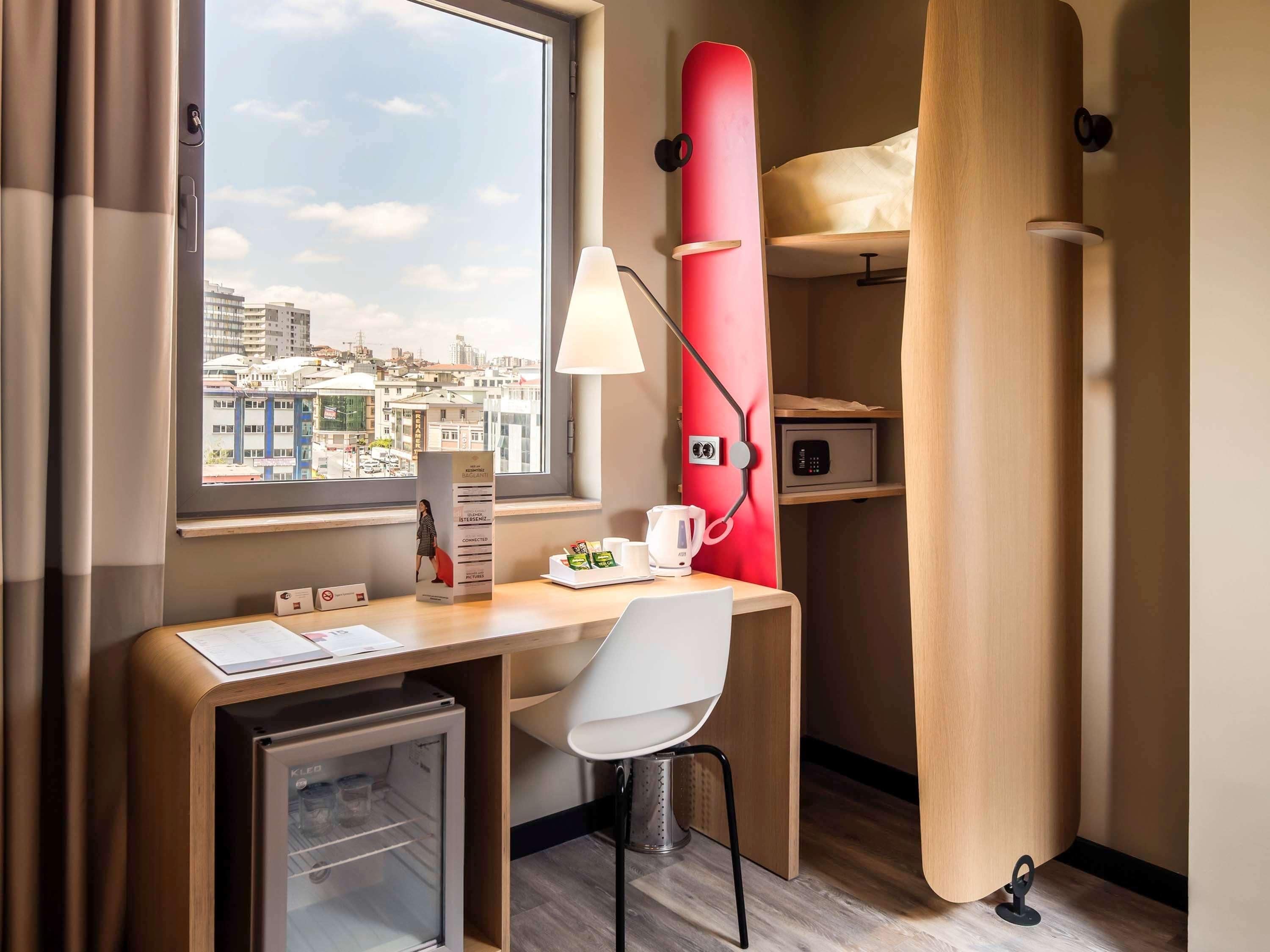 Habitación de hotel compacta con escritorio, silla, mini refrigerador, unidad de almacenamiento roja y vista panorámica de la ciudad.