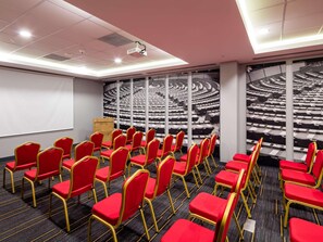 Un salón de conferencias con filas de sillas rojas, un proyector y una pantalla grande.