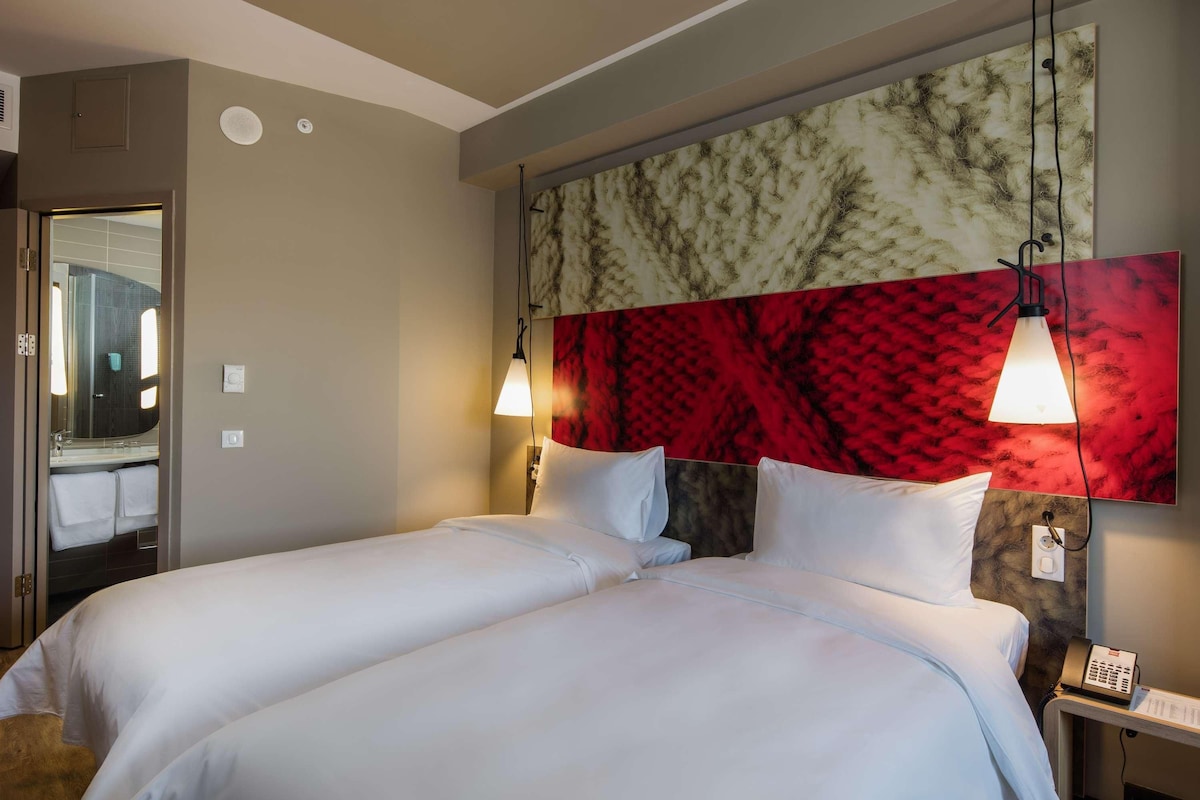 A hotel room with a bed, two bedside tables, a nightstand, a mirror, and a red and white abstract wall art.