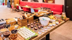 Un bufé con diversos alimentos y bebidas, un televisor montado en la pared y una zona de estar con sillones tapizados en rojo.
