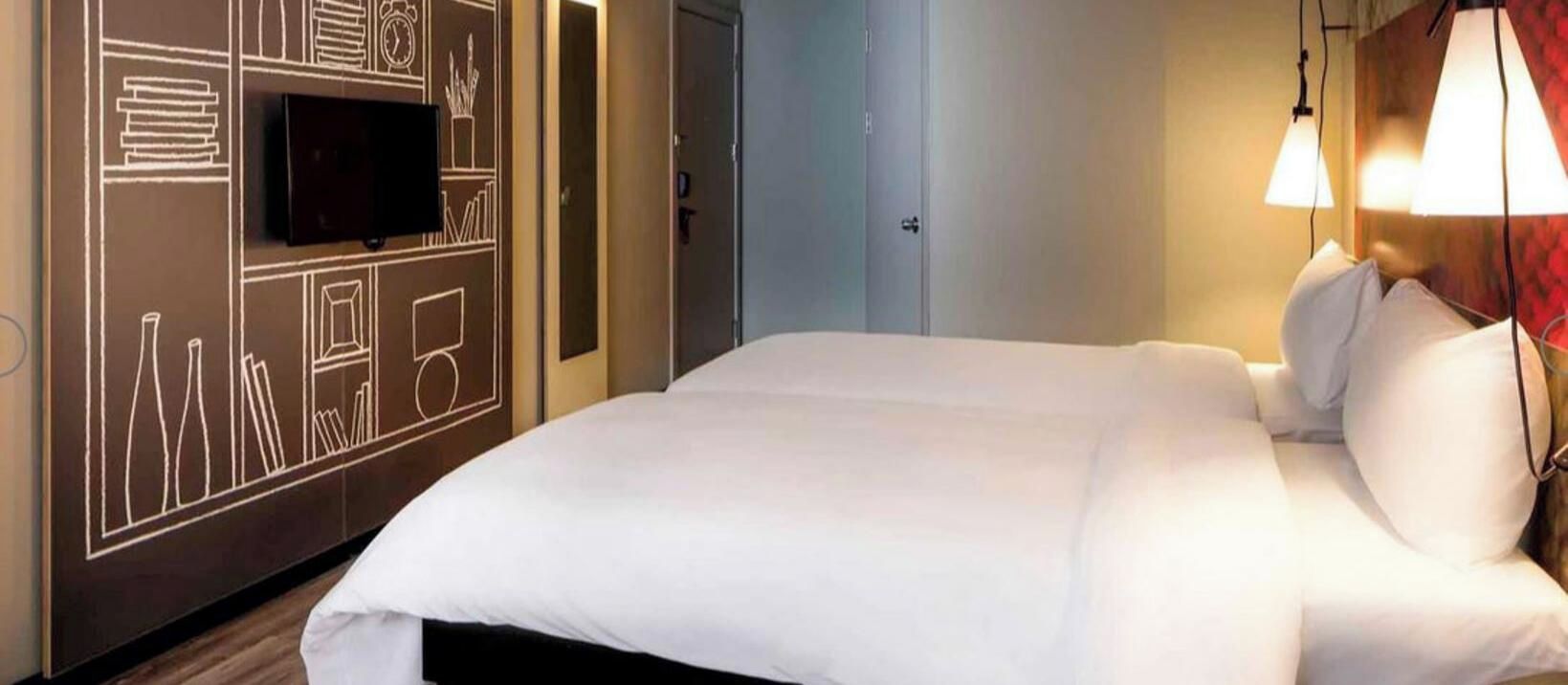 Hotellihuone, jossa on sänky, seinälle asennettu taulutelevisio ja ovi.