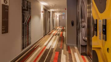 A hotel corridor with a striped carpet, elevator, and a wall-mounted sign indicating room numbers.