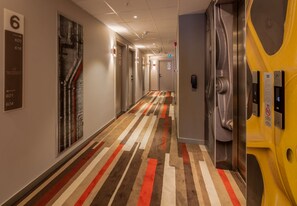Pasillo de hotel con alfombra a rayas, un ascensor y un letrero en la pared con los números de las habitaciones.