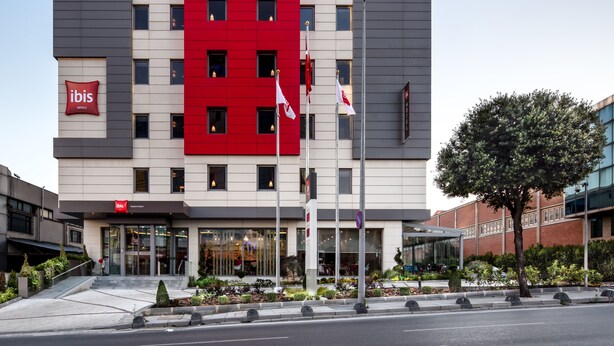 A modern hotel building with a prominent red and grey facade, multiple windows, and a visible entrance. - Ibis Istanbul West (Istanbul)
