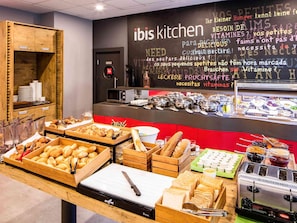 Buffet montado com diversos alimentos, área de cozinha e parede com texto em vários idiomas.