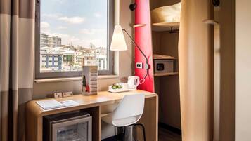 A compact hotel room with a desk, chair, mini-fridge, red storage unit, and a view of the cityscape.