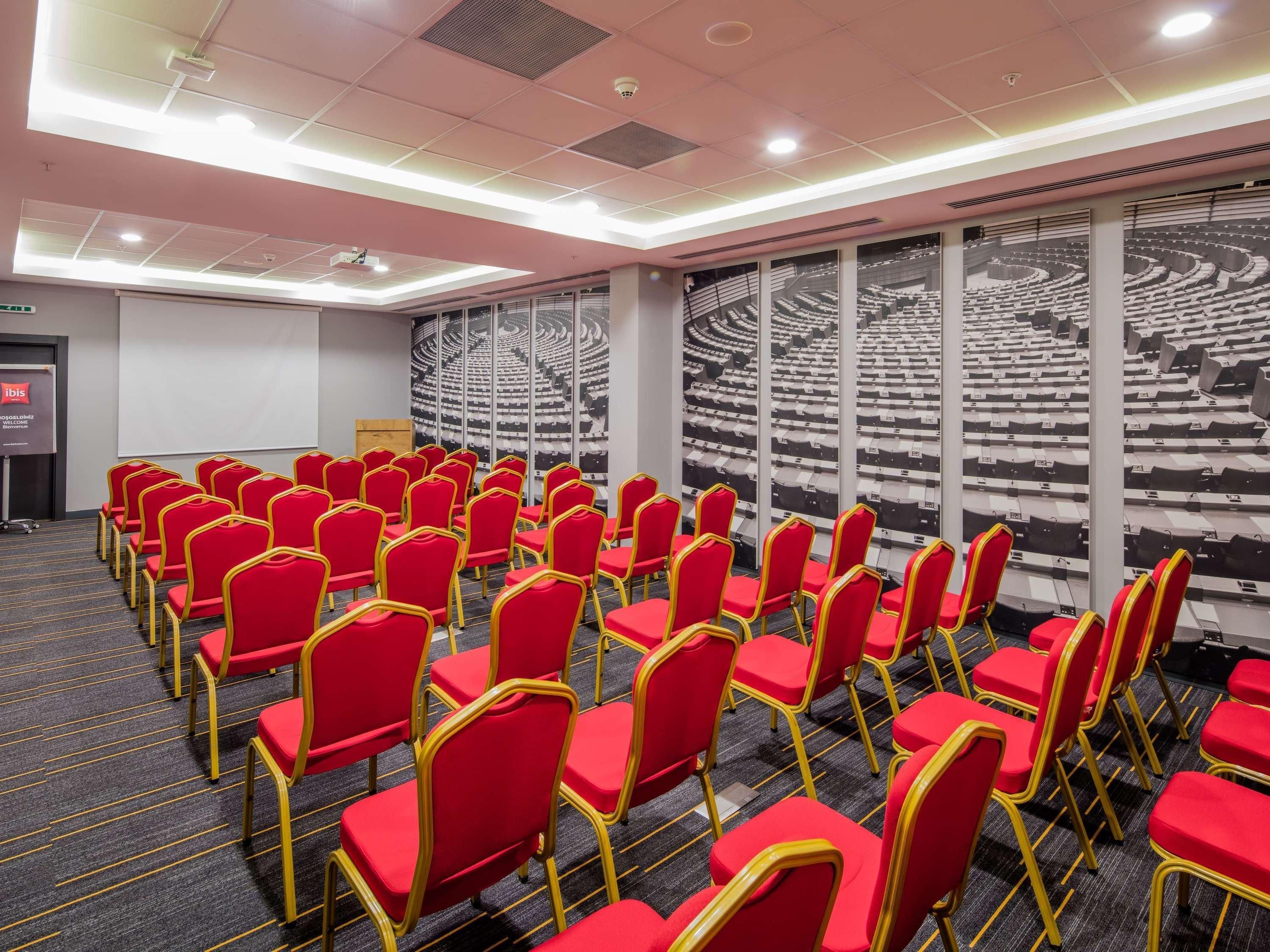 A conference room with rows of red chairs, a large screen, and a mural of a stadium.