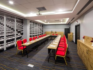 Een congreszaal met een lange tafel, rode stoelen, een beamer en een groot zwart-wit schilderij.