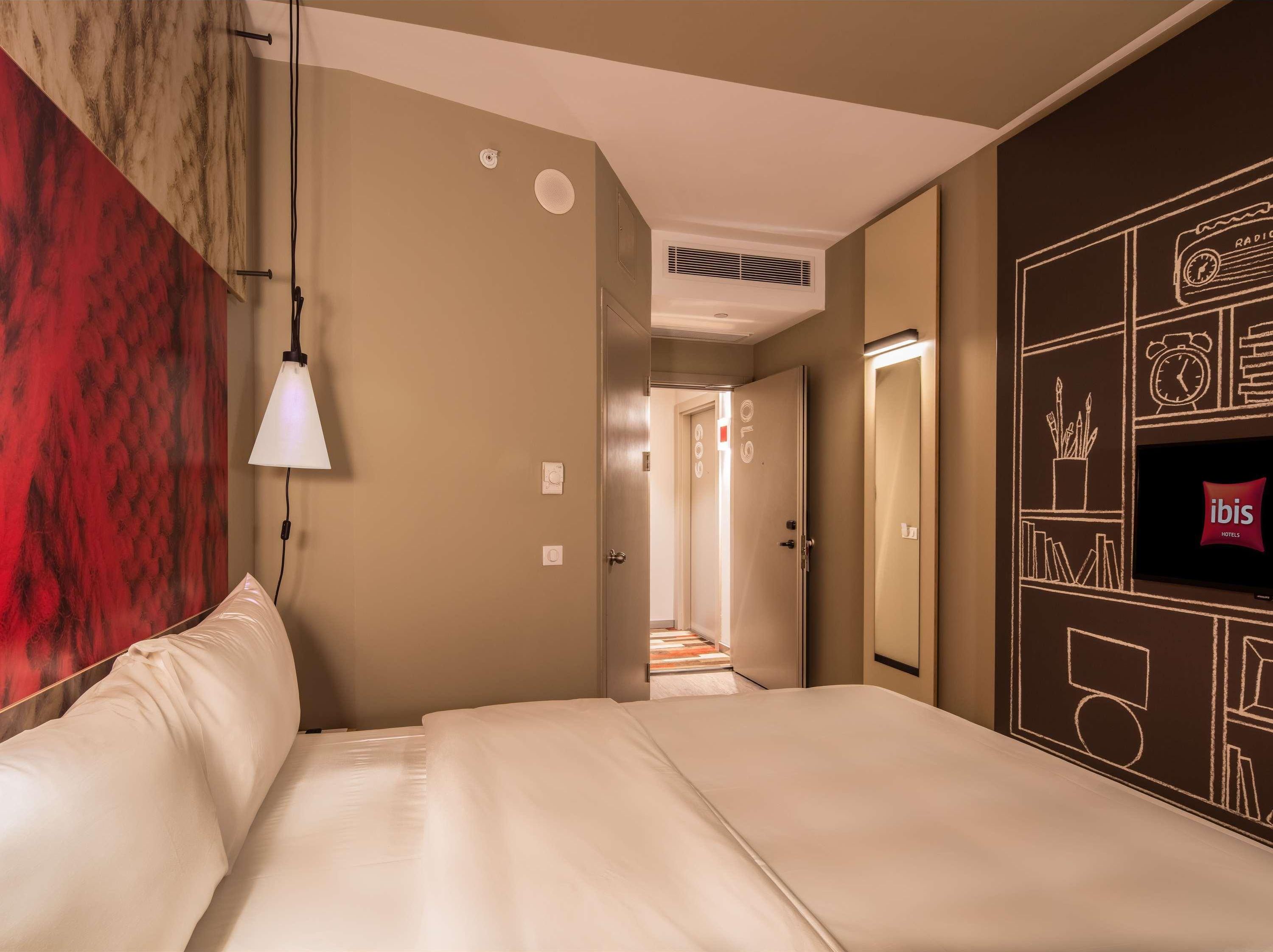 A hotel room with a bed, a wall-mounted TV, a red textured wall, and a hallway leading to another room.