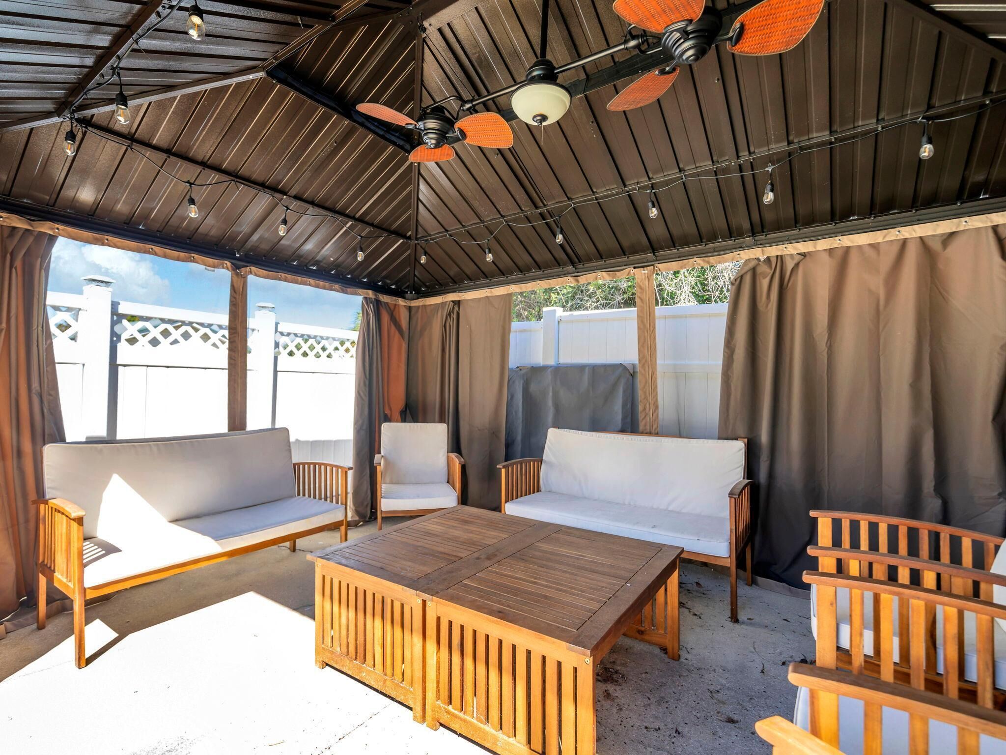 An outdoor patio with wooden furniture, including sofas and a table, under a metal roof with ceiling fans.