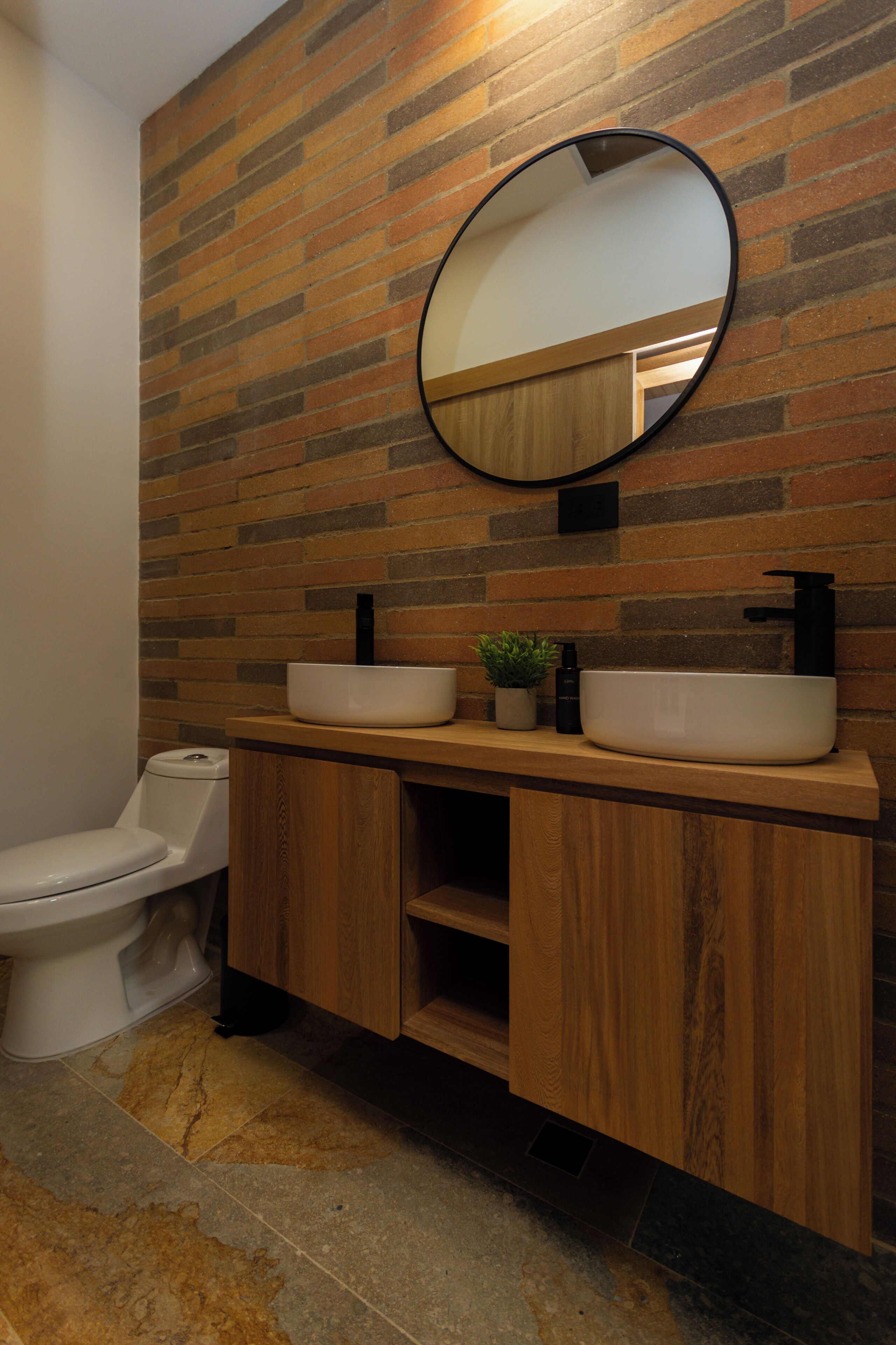 A bathroom with a wooden vanity, two sinks, a toilet, and a mirror on a brick wall.