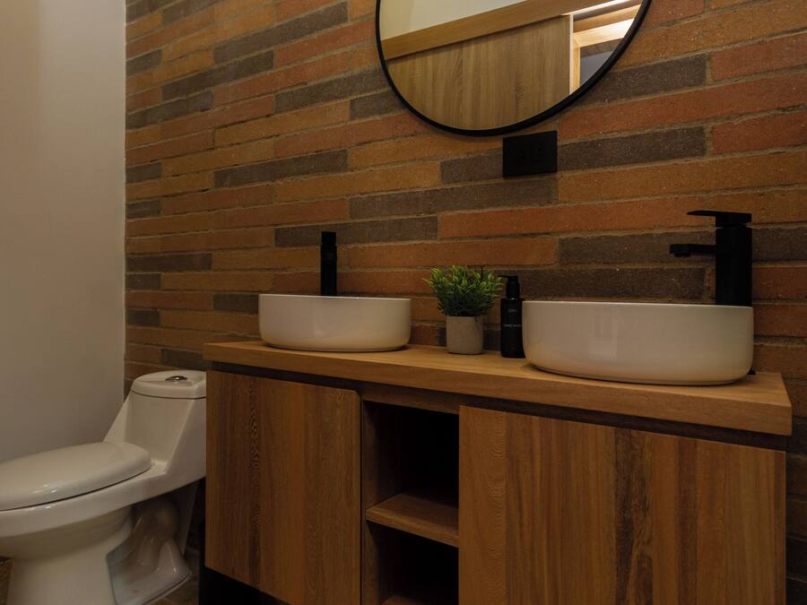A bathroom with a wooden vanity, two sinks, a toilet, and a mirror on a brick wall.