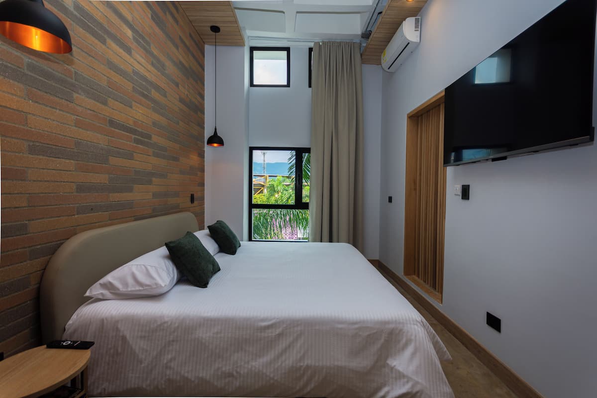 Ein Hotelzimmer mit Backsteinwand, einem großen Bett mit Kissen, einem an der Wand montierten Fernseher und einem Fenster mit Blick ins Grüne.
