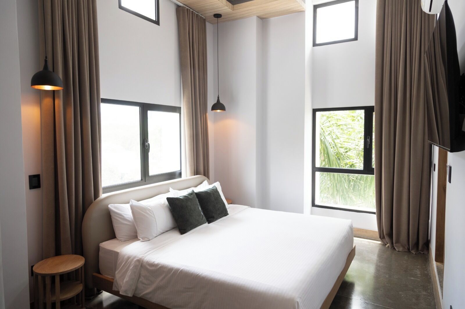 Un dormitorio con cama, dos ventanas, un televisor montado en la pared y una mesita.