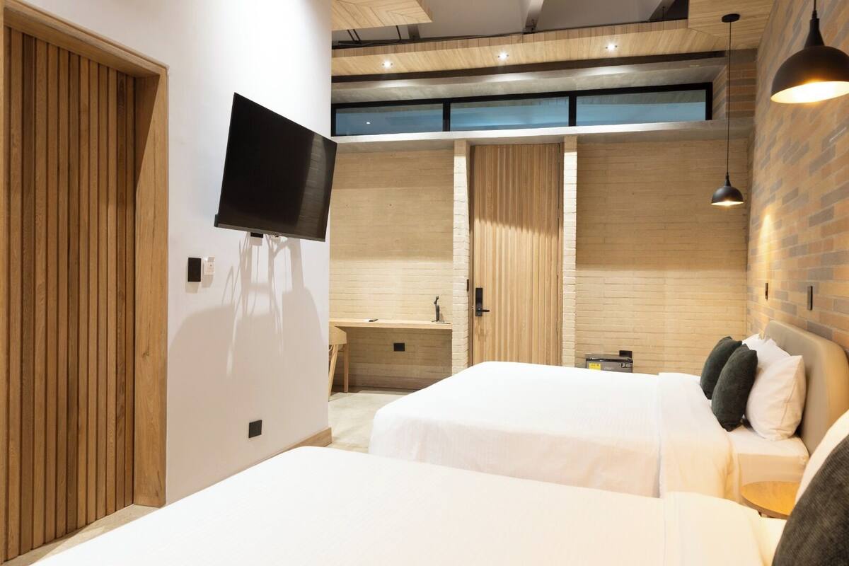 Ein modernes Hotelzimmer mit zwei Betten, einem Fernseher, einem Schreibtisch und einem Badezimmer mit Holztür.