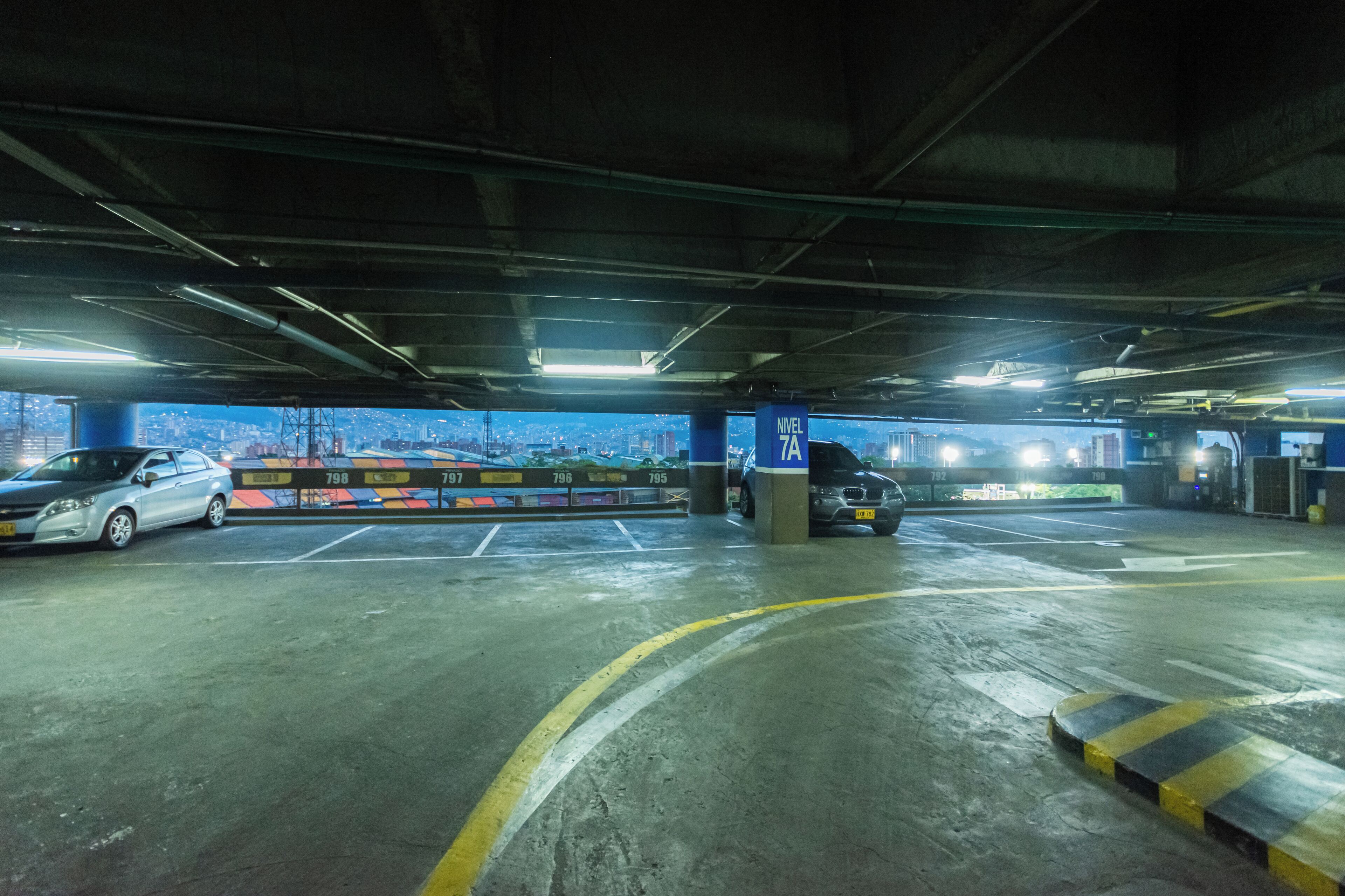 Un aparcamiento subterráneo con varios niveles, plazas de aparcamiento delimitadas y vistas a un paisaje urbano a través del techo abierto.