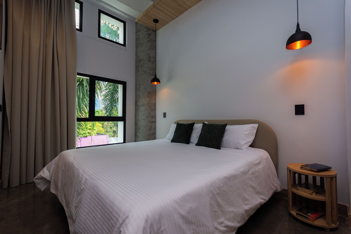 A bedroom with a large bed, a window with a view of greenery, and a small wooden side table with books.