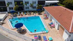 Una piscina con sillas de descanso y sombrillas, rodeada por un edificio con tejado de baldosas y una zona vallada.