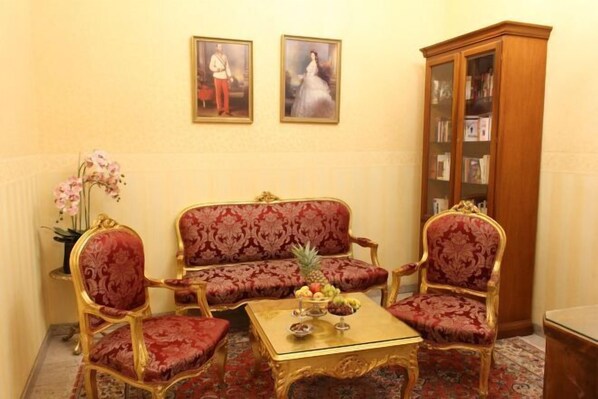 Ein Zimmer mit einem roten, bezogenen Sofa, zwei passenden Sesseln, einem kleinen Couchtisch mit Obst, einem Bücherregal und einer Zimmerpflanze.