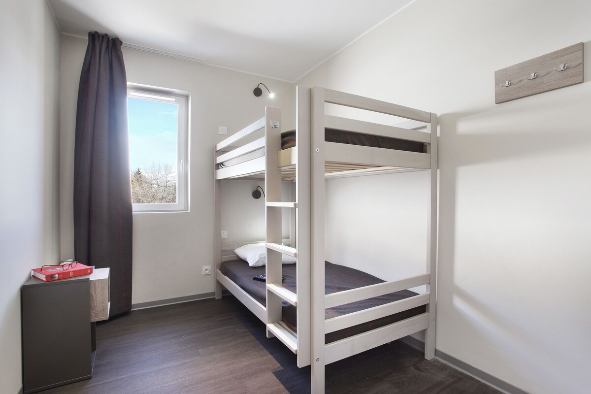 Une chambre avec un lit superposé, une fenêtre, un bureau et un luminaire fixé au mur.