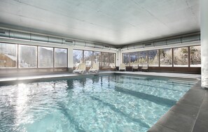 Una piscina interior con agua cristalina, sillones blancos para relajarse y grandes ventanales con vistas a la montaña.