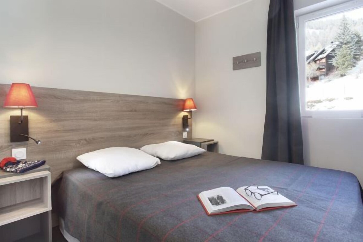 Una habitación de hotel con una cama, dos lámparas de noche, una mesita de noche, un ventanal con cortinas y vistas a un paisaje nevado.