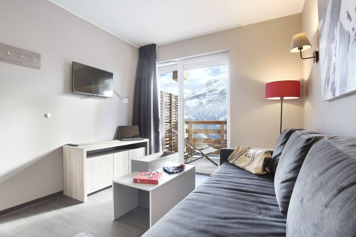 Una sala de estar moderna con un sofá, un televisor de pantalla plana, un escritorio con una computadora portátil, una lámpara y vistas a montañas a través de un amplio ventanal.
