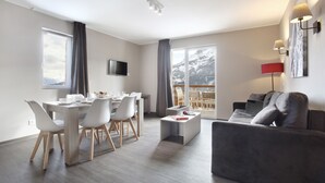 A modern living room with a grey sofa, a dining area with a table and chairs, and a large window with a view of snow-capped mountains. - Residence Odalys Le Village de Praroustan (Uvernet-Fours)