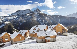Un grupo de cabañas de madera con tejados cubiertos de nieve, frente a montañas nevadas y cielos despejados.