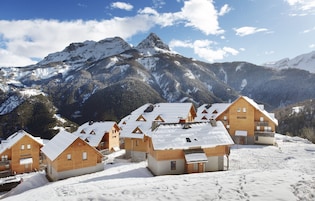 Um conjunto de cabanas de madeira com telhados cobertos de neve, com montanhas nevadas e céu claro ao fundo.