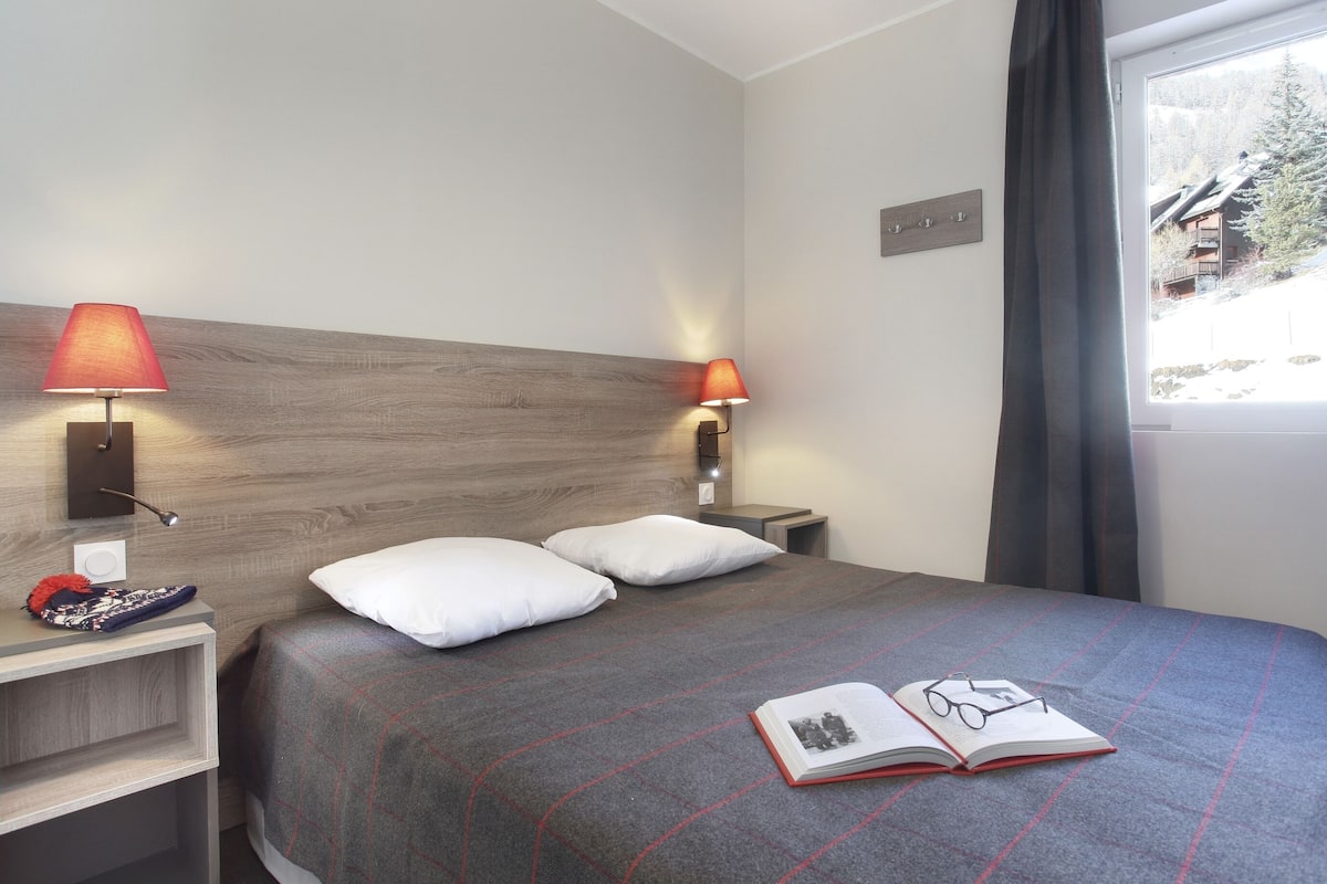 Habitación de hotel con cama, dos lámparas de noche, una mesita de noche, una ventana que ofrece vistas a un edificio y un libro sobre la cama.