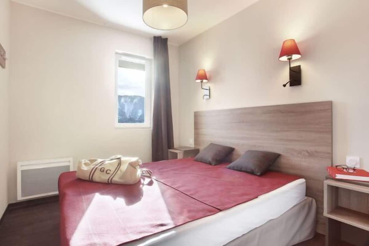 Habitación de hotel con cama, dos lámparas de noche, ventana con cortinas y mesita de noche con lámpara.