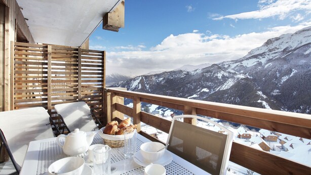 A balcony with a table set for breakfast, wooden railings, and a mountain view. - Residence Odalys Le Village de Praroustan (Uvernet-Fours)