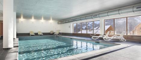 An indoor swimming pool with clear water, white lounge chairs, and large windows.