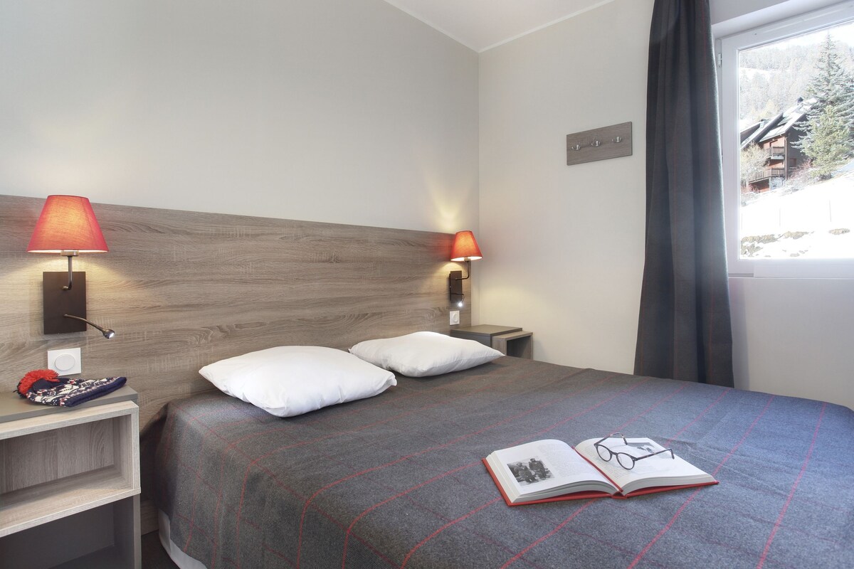 Une chambre d’hôtel avec un lit, deux lampes de chevet, une table de nuit, une fenêtre donnant sur un immeuble et un livre posé sur le lit.