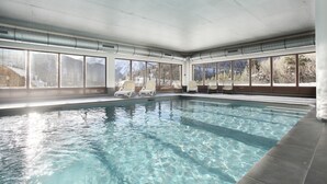 An indoor swimming pool with clear water, white lounge chairs, and large windows offering mountain views. - Residence Odalys Le Village de Praroustan (Uvernet-Fours)