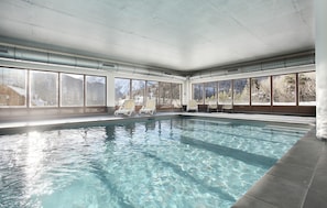 An indoor swimming pool with clear water, white lounge chairs, and large windows offering mountain views.
