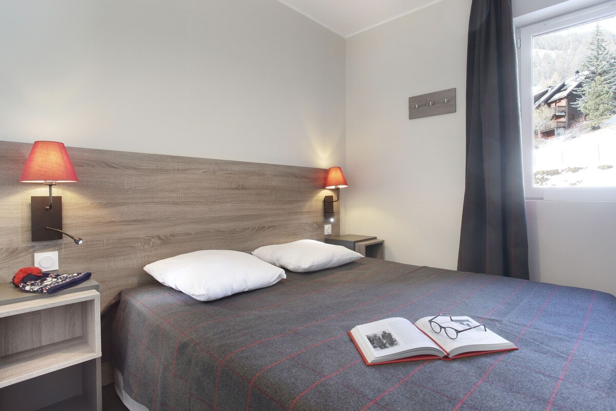 Un lit bien fait, recouvert d’une couverture à carreaux rouges et gris, avec deux oreillers blancs et un livre ouvert posé dessus, accompagné d’une paire de lunettes. On y trouve deux lampes de chevet à abat-jour rouge, une tête de lit en bois et une fenêtre donnant sur un bâtiment et des arbres.