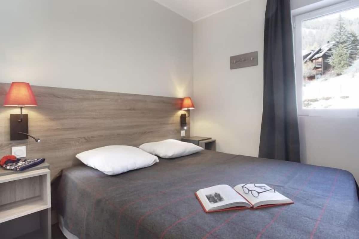 Una habitación de hotel con una cama, dos lámparas de noche, una mesita de noche, un ventanal con cortinas y vistas a un paisaje nevado.