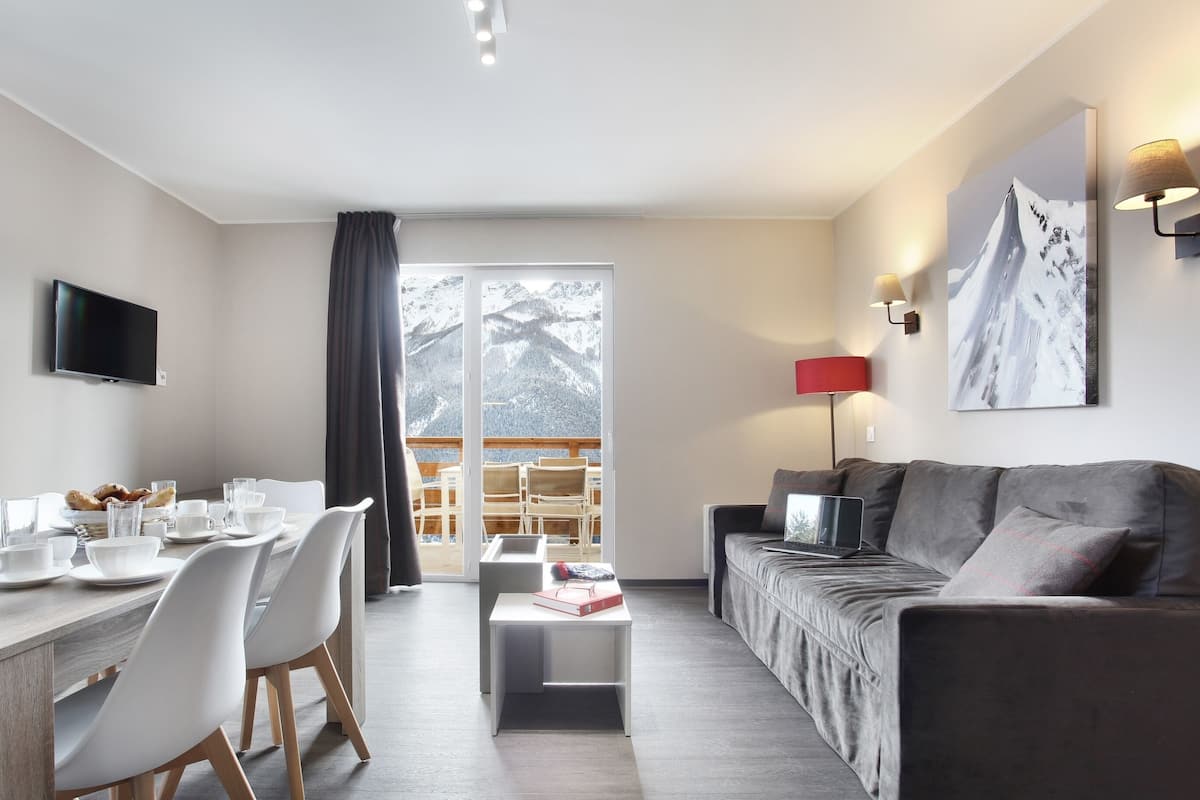 Una sala de estar moderna con comedor, sof á, televisor y balcón con vistas a montañas nevadas.
