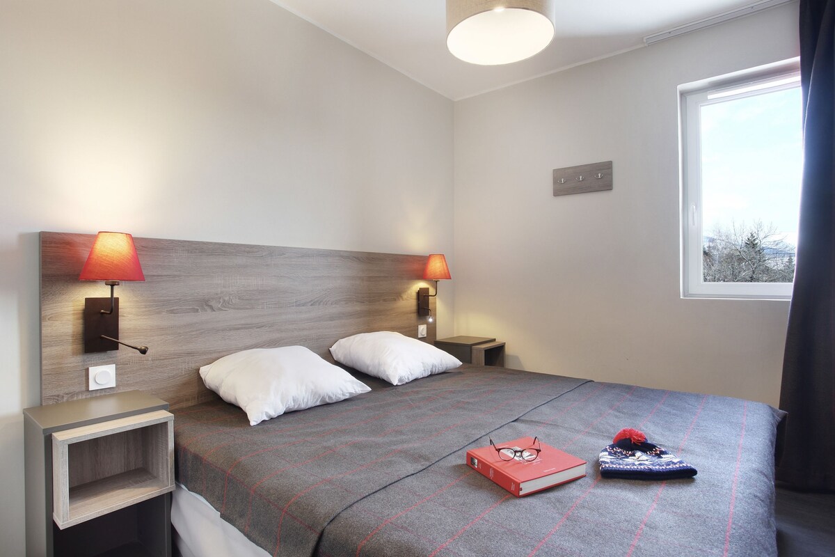 Un lit bien fait, avec une couverture à carreaux, deux lampes de chevet, un livre et des chaussons posés sur le lit.