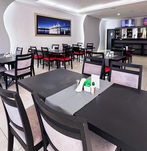 A modern hotel dining area with multiple tables and chairs, a large painting on the wall, and a bar area in the background. - Bella Vista Hotel (Encarnacion)