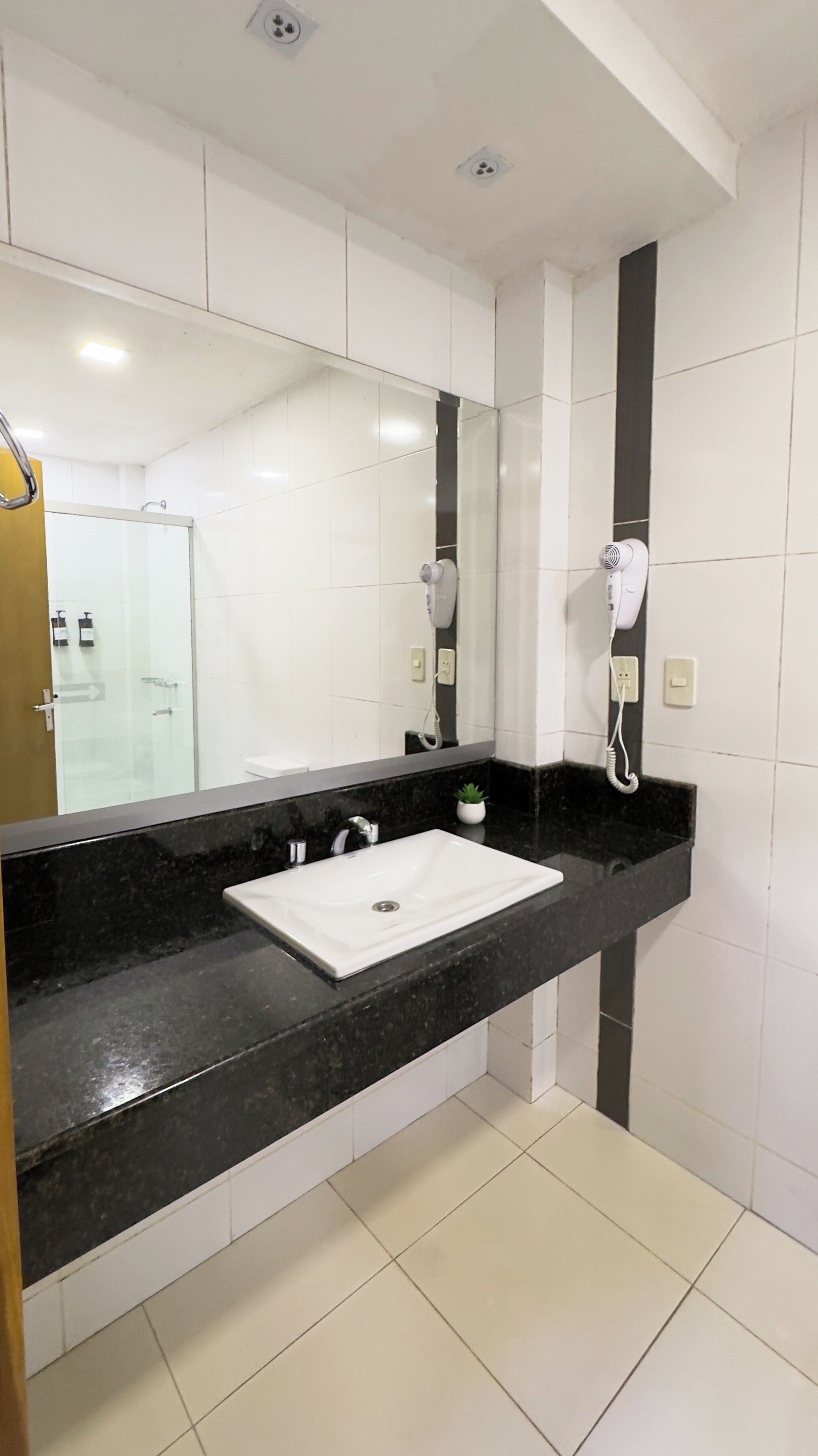 Un baño con lavamanos blanco, encimera negra y un espejo grande. Hay secadores de pelo montados en la pared.