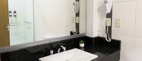 Un baño con lavamanos blanco, encimera negra y un espejo grande. Hay secadores de pelo montados en la pared.