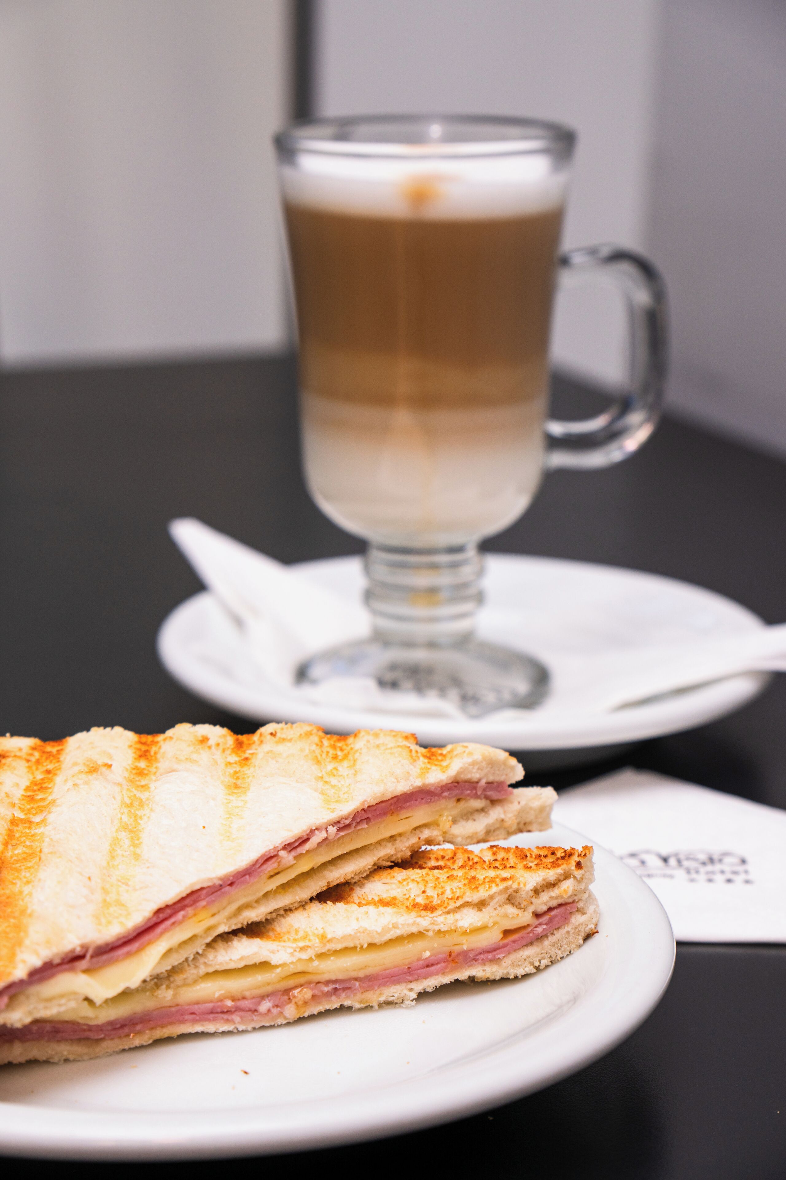 A sandwich on a plate, a glass of coffee, and a napkin on a table.