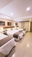 A hotel room with four beds, a wooden headboard, a large window with curtains, and a door.